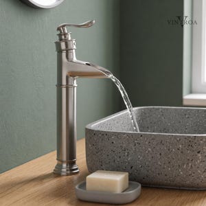 May include: A brushed nickel bathroom faucet with a waterfall spout, dispensing water into a gray speckled sink. A bar of soap rests on a small gray soap dish. The faucet is mounted on a wooden surface.