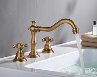 Op de afbeelding: Een messing kraan in vintage-stijl met een gebogen uitloop en kruisvormige knoppen. De kraan is gemonteerd op een wit oppervlak, met water dat in een witte wastafel stroomt. Het ontwerp omvat een centrale kraan en twee afzonderlijke handgrepen.