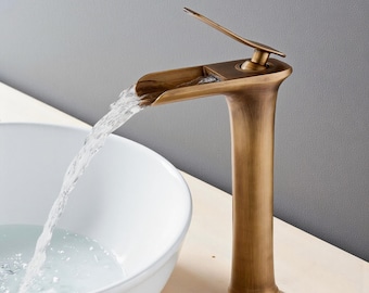 Op de afbeelding: Een bronzen badkamerkraan met een watervaluitloop. Water stroomt uit de uitloop in een witte keramische wastafel. De kraan heeft een hoge, gebogen behuizing en een hendel. De merknaam "VINTROA" is zichtbaar.