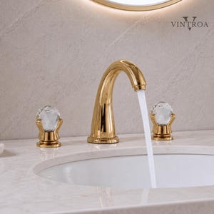 May include: A gold-colored bathroom faucet with a curved spout and two crystal ball handles. The faucet is turned on, and water is flowing into a white sink. The faucet has a decorative design and is set against a light-colored background.