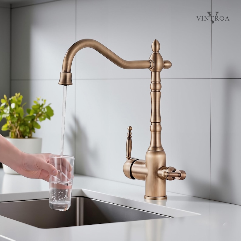 Puede incluir: Grifo de cocina de tono bronce con un ca&ntilde;o curvo y detalles decorativos. El agua fluye hacia un vaso transparente. El grifo tiene un dise&ntilde;o vintage, con una palanca y dos manijas. El grifo est&aacute; montado en una encimera blanca.