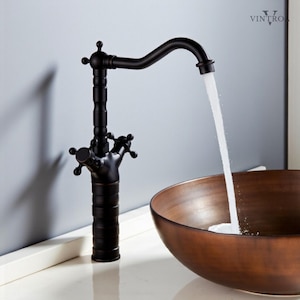 Puede incluir: Un grifo de baño negro de estilo vintage con un caño curvo, agua fluyendo hacia un cuenco de color cobre. El grifo tiene manijas en forma de cruz y detalles decorativos. El grifo está sobre una superficie blanca.
