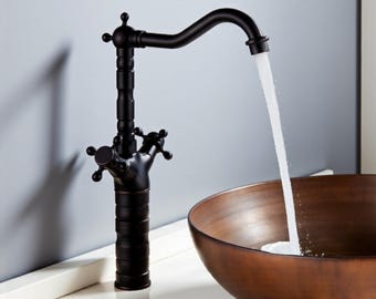 Op de afbeelding: Een zwarte badkamerkraan in vintage-stijl met een gebogen uitloop, water stroomt in een koperkleurige kom. De kraan heeft kruisvormige handgrepen en decoratieve details. De kraan staat op een wit oppervlak.