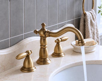 May include: A brushed brass bathroom faucet with a curved spout and two lever handles, mounted on a beige marble countertop. Water flows from the spout into a white sink basin. The VINTROA logo is visible on the gray tiled wall behind.