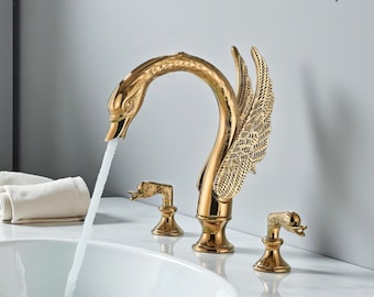 Op de afbeelding: Een goudkleurige zwaanvormige kraan met stromend water. De kraan heeft ingewikkelde details, waaronder een zwanenkop en gevederde vleugels. Twee kleinere zwaanvormige hendels zijn ook zichtbaar. De merknaam "VINTROA" staat rechtsboven.