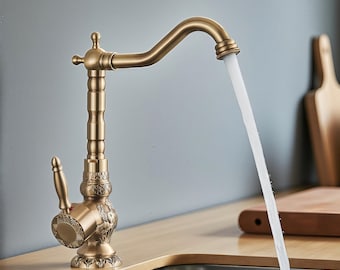 May include: A bronze-colored kitchen faucet with a curved spout and ornate detailing. Water flows from the faucet into a stainless steel sink. The faucet features a decorative handle and base, adding a vintage touch to the kitchen.