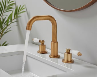 Op de afbeelding: Een goudkleurige badkamerkraan met een gebogen uitloop en twee aparte handgrepen. De handgrepen hebben witte accenten. Water stroomt uit de kraan in een witte wastafel. Een ronde spiegel staat op de achtergrond.