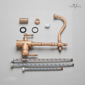 Puede incluir: Un grifo de cocina de bronce de estilo antiguo, con varios componentes dispuestos alrededor. El grifo presenta un ca&ntilde;o curvo, una manija de palanca y detalles decorativos. Incluye l&iacute;neas de suministro flexibles y otras piezas.