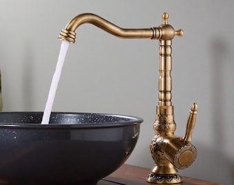 Op de afbeelding: Een koperkleurige kraan in antieke stijl met gedetailleerde bloemgravures en een hendel, waaruit een constante waterstraal in een donkere, gespikkelde waskom stroomt. Het VINTROA-logo is zichtbaar in de rechterbovenhoek van de afbeelding.