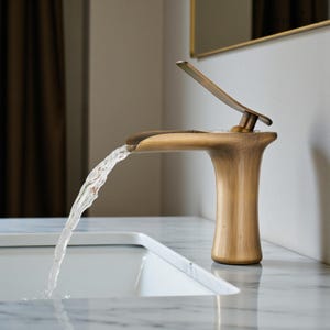 Puede incluir: Un grifo de color bronce con agua que fluye hacia un lavabo blanco. El grifo tiene un diseño curvo único con una palanca en la parte superior. El accesorio está colocado contra una pared de tonos neutros y una encimera de mármol.