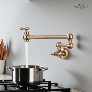 Op de afbeelding: Een aan de muur gemonteerde, messingkleurige potvuller-kraan met een waterstraal die in een roestvrijstalen pot op een fornuis stroomt. De kraan heeft een vintage design en is op een lichtgrijze muur gemonteerd. De merknaam "VINTROA" is zichtbaar.