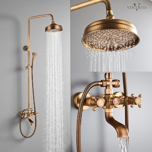 May include: Antique brass shower fixture with a round showerhead and handheld sprayer. The showerhead is positioned above the faucet and water is flowing. The fixture has a vintage design with cross-shaped handles and a curved spout.