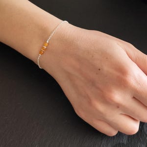 Op de afbeelding: Een verfijnde zilverkleurige kettingarmband met drie kleine, ronde, doorschijnende oranje kralen in het midden. De armband is gemaakt van een fijne metalen schakelketting en bevat kleine zilveren tussenstukjes tussen de stenen.