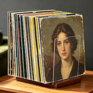 Könnte beinhalten: Ein Schallplattenständer aus Holz mit einer Sammlung von Schallplatten. Die vordere Schallplatte zeigt das Porträt einer Person mit braunen Haaren. Der Ständer hat einen transparenten Acrylteiler und einen Sockel aus dunklem Holz.