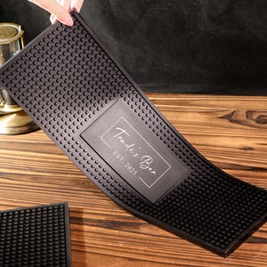 Può includere: Tappetino da bar in gomma nera con superficie strutturata e inserto rettangolare. L'inserto presenta il testo "Trude's Bar EST. 2025" in bianco, con un carattere elegante. È visibile anche un tappetino più piccolo e simile.