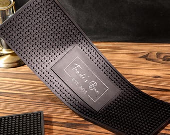 May include: Black rubber bar mat with a textured surface and a rectangular inset. The inset features the text "Trude's Bar EST. 2025" in a white, elegant font. A smaller, similar mat is also visible.