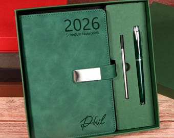 Puede incluir: Un juego de regalo verde que incluye un cuaderno de programación de 2026, un bolígrafo y un lápiz óptico. El cuaderno tiene la inscripción "2026 Schedule Notebook" y el nombre "Phil".