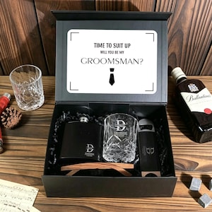 May include: A black gift box containing a hip flask, a rocks glass, a bottle opener, and wooden sunglasses. The box lid has the text "TIME TO SUIT UP WILL YOU BE MY GROOMSMAN?" A bottle of Ballantine's whisky is visible.