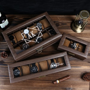 May include: Wooden watch display cases with glass tops. The cases are brown with individual compartments lined with tan fabric. One case has the initials "AK" and another has the name "Matthew" etched on the glass. Several watches are displayed.