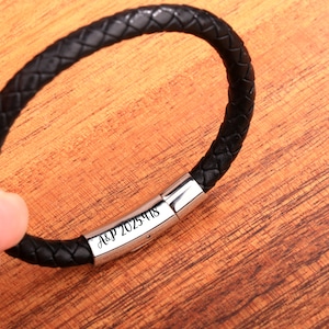 May include: Black braided leather bracelet with a silver clasp. The clasp is engraved with the text "A&P 2025-418". The bracelet is shown on a wooden surface.