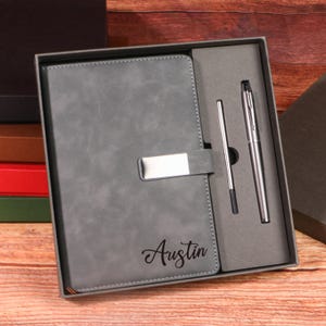 Puede incluir: Un juego de regalo que incluye un cuaderno de cuero gris con cierre plateado y el nombre "Austin" en cursiva. Viene con un bolígrafo plateado y una recarga, todo en una caja.