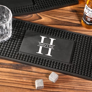 May include: A black bar mat with a textured surface and the text "II Harry's Bar II" in white. A clear glass, a bottle of beer, and two grey stone cubes are also visible on a wooden surface.