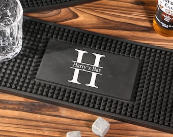 May include: A black bar mat with a textured surface and the text "II Harry's Bar II" in white. A clear glass, a bottle of beer, and two gray stone cubes are also visible on a wooden surface.