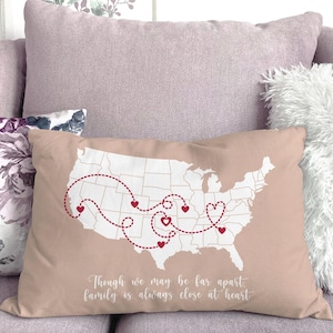 May include: A rectangular pillow with a map of the United States, featuring red heart accents and dotted lines connecting various locations. The pillow has the text "Though we may be far apart, family is always close at heart". The pillow is a light tan color.
