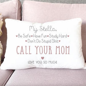 May include: White rectangular pillow with the text "My Stella, Be Safe, Have Fun, Study Hard, Don't Do Stupid Shit, CALL YOUR MOM, LOVE YOU SO MUCH!" in a mix of script and block letters. The pillow is on a pink couch.