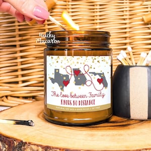 May include: A brown glass jar candle with a label that reads "The love between Family KNOWS NO DISTANCE." The label features gray state outlines with red hearts. A match is being lit, and a black and gray match holder is next to the candle.