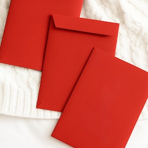 May include: Three red envelopes are arranged on a white, textured surface. The envelopes are rectangular with a flap closure. The vibrant red color of the envelopes contrasts with the white background, creating a visually appealing composition.
