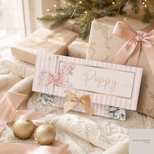 Puede incluir: Una tarjeta rectangular con un patrón de rayas rosas y blancas, una ilustración de un caballo de carrusel dorado y el nombre Poppy escrito en letra dorada. La tarjeta está decorada con un lazo de satén dorado y un borde rectangular.