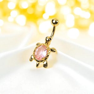 Puede incluir: Un piercing para el ombligo de color dorado con un diseño de tortuga. La tortuga tiene una gema rosa de forma ovalada en su caparazón. El anillo tiene un diseño de barra curva con una pequeña bola dorada en la parte superior.