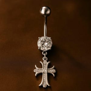 May include: A silver belly button ring featuring a large, clear, round gemstone and a dangling cross pendant. The cross is embellished with smaller, sparkling stones. The top of the ring has a small, round silver ball.