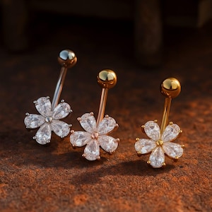 Puede incluir: Tres piercings para el ombligo con diseños de flores. Las flores tienen piedras claras y brillantes y están engastadas en metal plateado, oro rosa y dorado. Las barras y las bolas también son plateadas, oro rosa y doradas. Las joyas se exhiben sobre una superficie marrón texturizada.