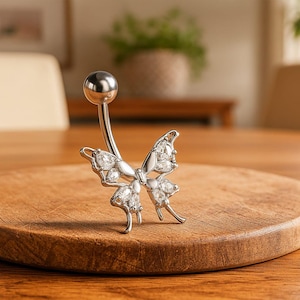 Puede incluir: Un piercing para el ombligo plateado con un diseño de mariposa. La mariposa tiene detalles claros y brillantes. El piercing se muestra sobre una tabla de cortar redonda de madera.