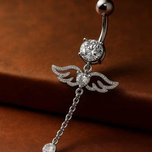 May include: Silver belly button ring with a dangling angel wing design. The ring features a curved barbell with a large, clear, round-cut crystal at the top and bottom. The angel wings are encrusted with smaller crystals.