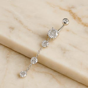 May include: A silver belly button ring with a curved barbell design. The ring features a small, round gem at the top and a dangling chain with four round, sparkling gems of varying sizes. The gems are clear and reflect light.