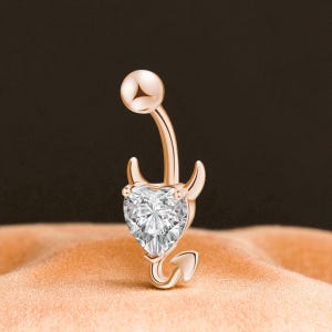 Può includere: Anello per ombelico in oro rosa con una gemma trasparente a forma di cuore. L'anello presenta un design a tema diavolo con corna e coda. La sfera superiore e la barra curva sono anch'esse in oro rosa.