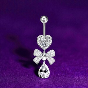 May include: A silver belly button ring featuring a heart-shaped top, a bow, and a teardrop-shaped bottom, all adorned with clear, sparkling stones. The background is a solid purple.