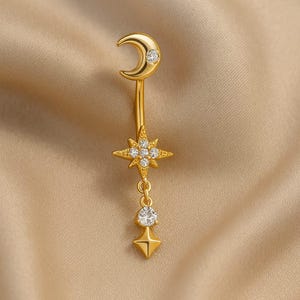 Puede incluir: Anillo para el ombligo de color dorado con una luna creciente con una gema transparente, una estrella con múltiples gemas transparentes y un dije colgante con una gema transparente y un dije cuadrado. La joya está sobre un fondo neutro.