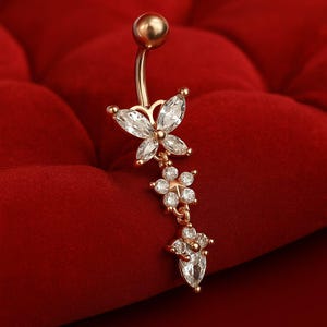 May include: A gold-colored belly button ring with a butterfly and floral design. The butterfly and flowers are adorned with clear, sparkling crystals. The curved barbell is topped with a gold ball.