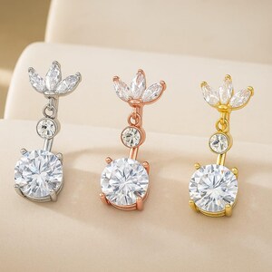Op de afbeelding: Drie navelpiercings in zilver, roségoud en goud. Elke ring heeft een grote, ronde, heldere edelsteen aan de onderkant, een kleinere edelsteen in het midden en een bloemmotief aan de bovenkant. De sieraden worden op een neutrale achtergrond gepresenteerd.