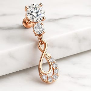 Op de afbeelding: Rosegouden oorbel met drie heldere, sprankelende zirkonia stenen. De bovenste steen is groot en rond, gevolgd door een kleinere ronde steen. Het onderste gedeelte heeft de vorm van een oneindigheidssymbool met kleinere stenen.