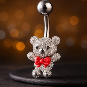 May include: A silver belly button ring featuring a sparkling teddy bear design. The bear is covered in clear crystals and has a bright red bow tie. The ring has a curved barbell with a silver ball at the top.