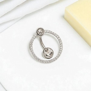 May include: A silver belly button ring with a curved barbell design. The top and bottom balls are clear, round gemstones. The ring features a circle of small, clear gemstones around the bottom ball. The jewelry is set against a white background.