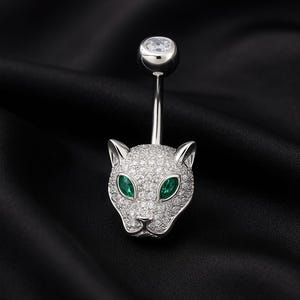 Könnte beinhalten: Ein silberner Bauchnabelpiercing mit einem Pantherkopf-Design. Der Pantherkopf ist mit kleinen, klaren Kristallen besetzt, mit smaragdgrünen Augen. Der obere Ball ist ein klarer Kristall. Der Schmuck ist vor einem schwarzen Hintergrund.