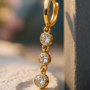 Könnte beinhalten: Ein goldfarbener Ohrring mit drei klaren, runden Edelsteinen, die an einem Bügel hängen. Die Edelsteine sind durch kleine goldene Ketten verbunden. Der Ohrring wird vor einem verschwommenen Hintergrund präsentiert, der das Funkeln des Schmucks hervorhebt.