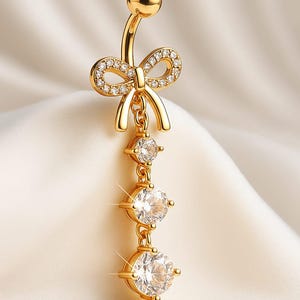 Puede incluir: Anillo para el ombligo dorado con un diseño de lazo incrustado con pequeños cristales transparentes. La parte colgante presenta tres cristales grandes, redondos y transparentes. La joya está sobre un fondo blanco suave.