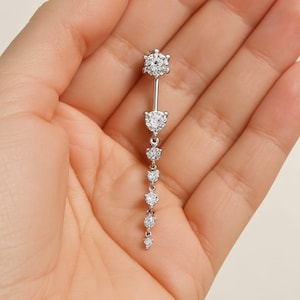 May include: A silver belly button ring with a series of clear gemstones. The top stone is a round cluster, followed by a heart-shaped stone, and then a line of round stones descending in size. The jewellery is held in a hand.